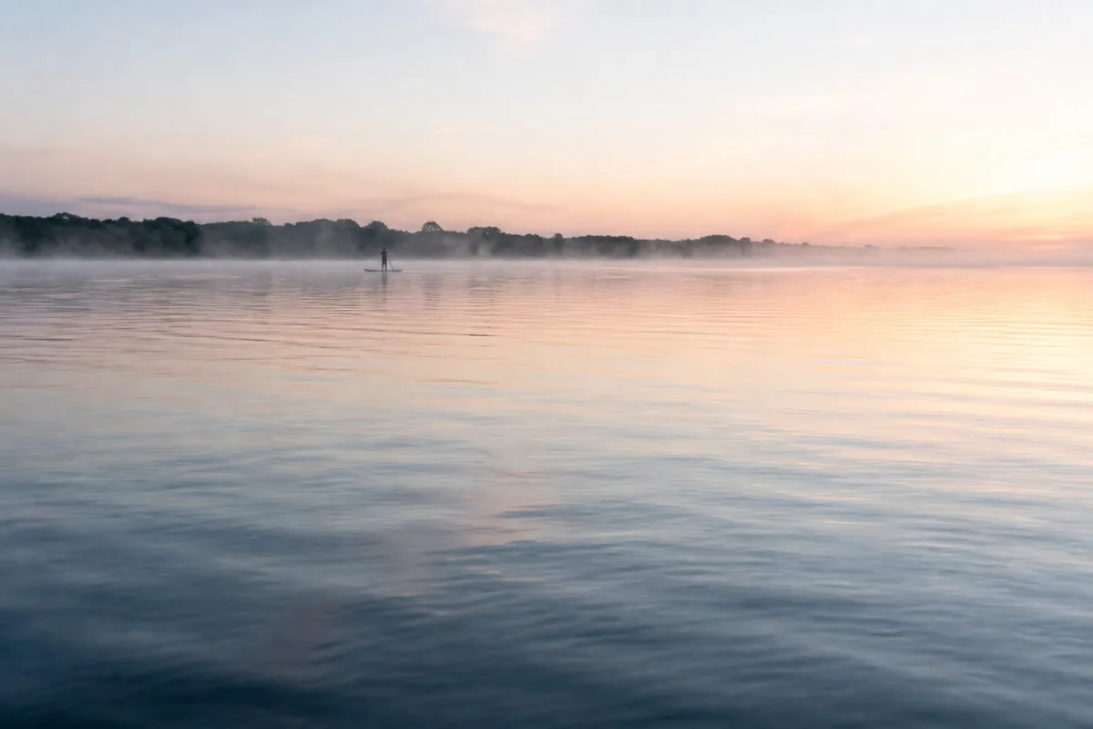 Plan d’eau calme au petit matin, silhouette lointaine debout sur une planche